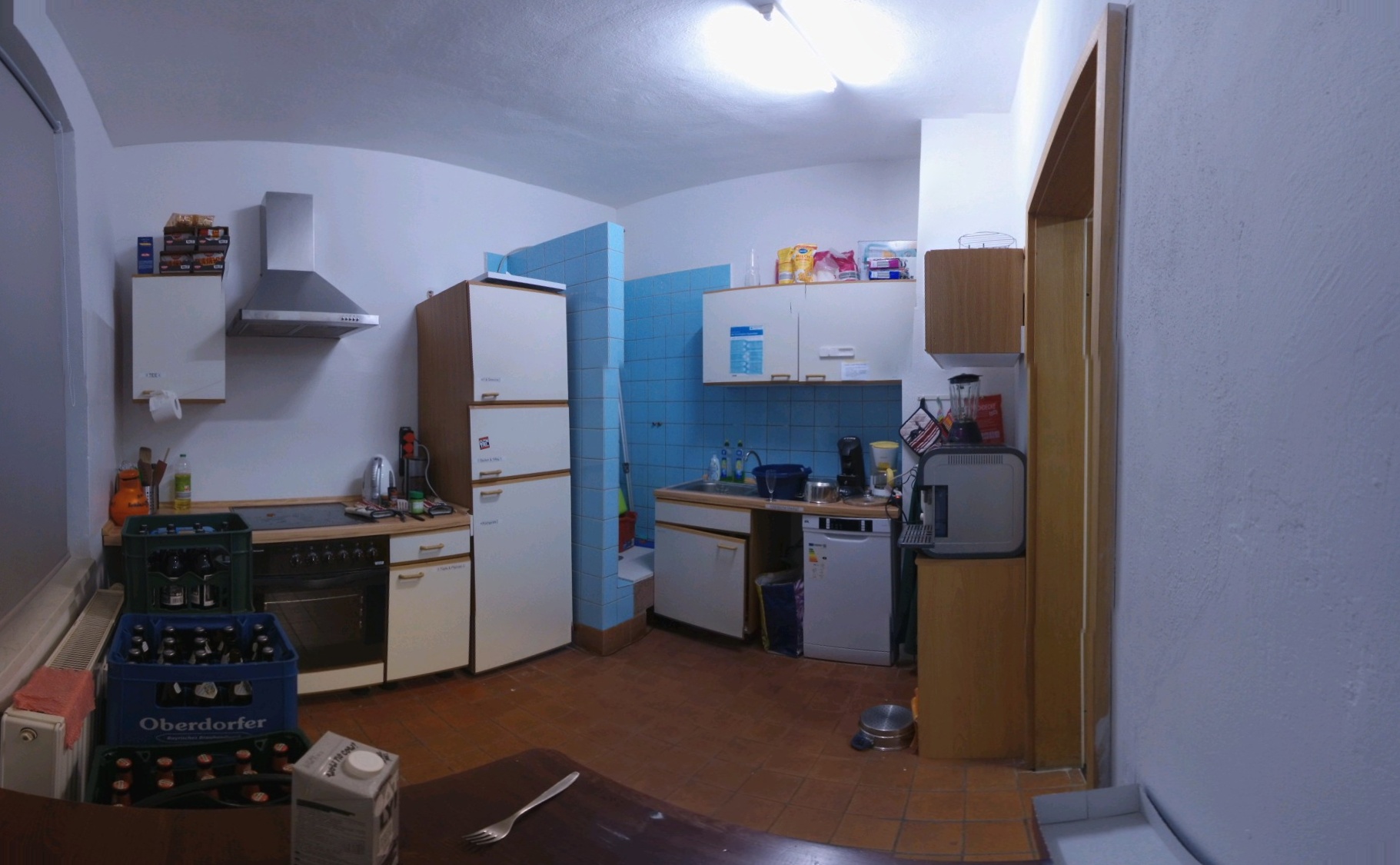 Eine unaufgeräumte Küche, welche einen blau gefließten Bereich enthält, welcher wie eine Dusche aussieht. Mitten in der Küche! Alles zum Kochen ist vorhanden, ein Nudelabgießtopf liegt auf dem Boden, eine Gabel und eine Packung Milchersatzflüssigkeit steht vor der Kamera und eine Toilettenpapierrolle wurde als Aufwischtücherersatz installiert